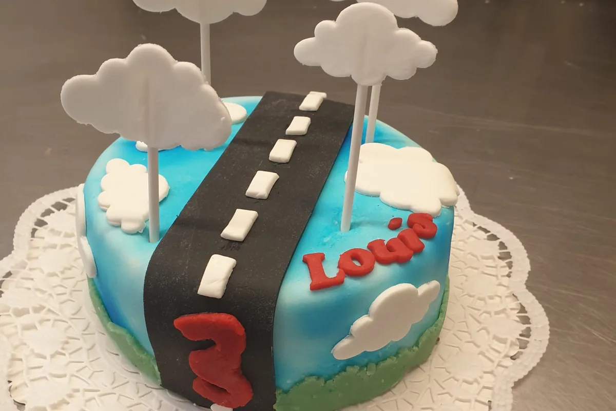 Eine Motivtorte zum dritten Geburtstag: eine Straße aus Fondant über blauem Himmel, darüber Wolken am Stiel.