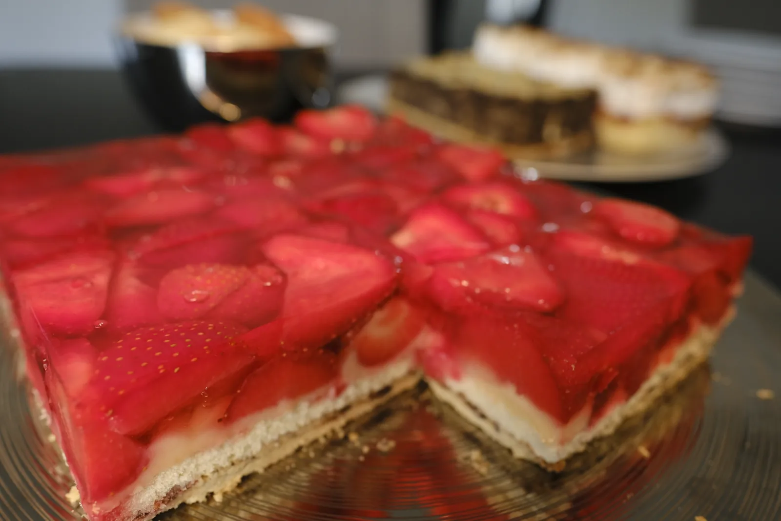 Erdbeer-Blechkuchen mit Guss, angeschnitten, dahinter weitere Kuchen.