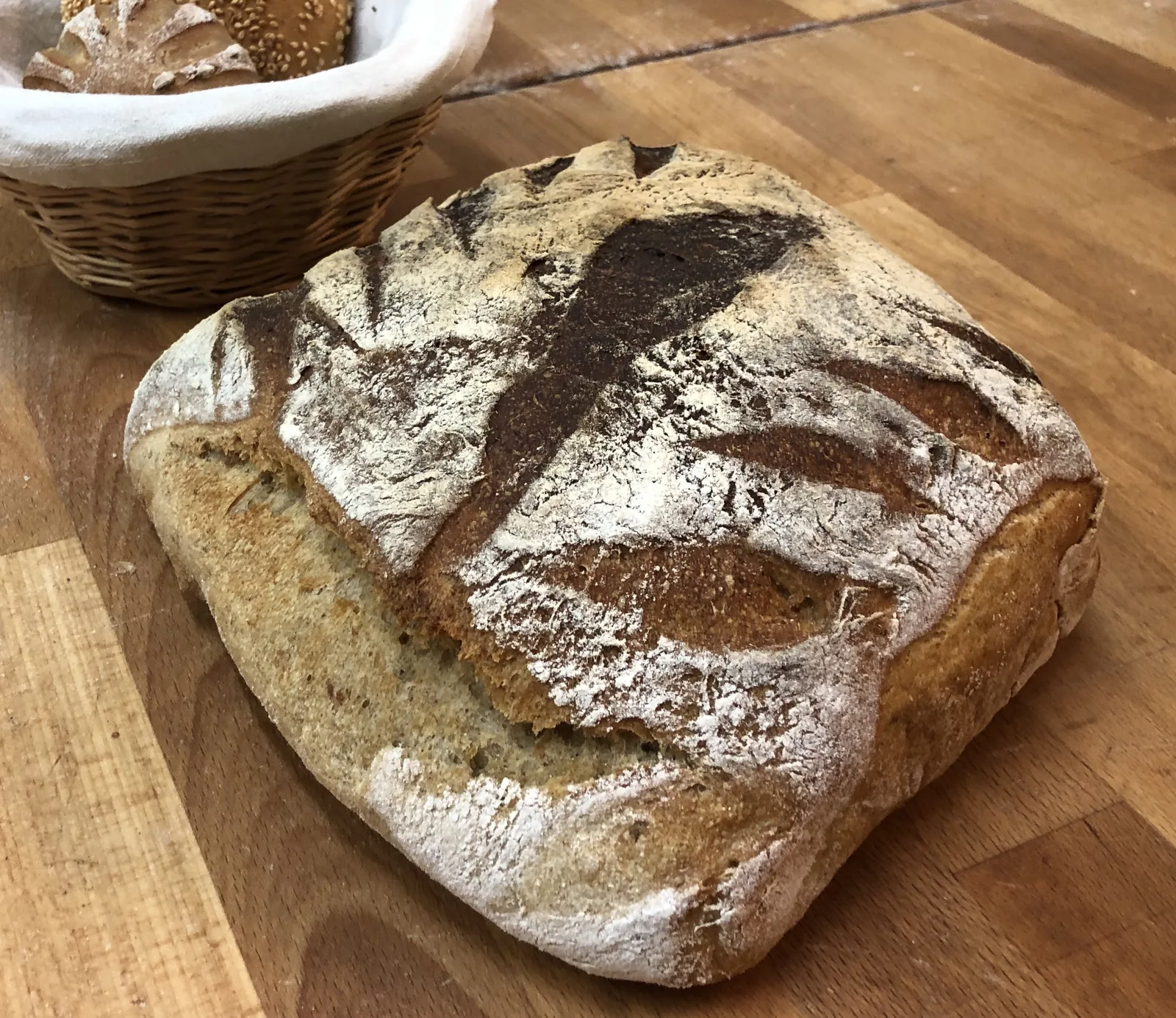 Ein runder Brotlaib mit hellem Mehlmuster auf einem Holzbrett, dahinter ein Gärkorb.