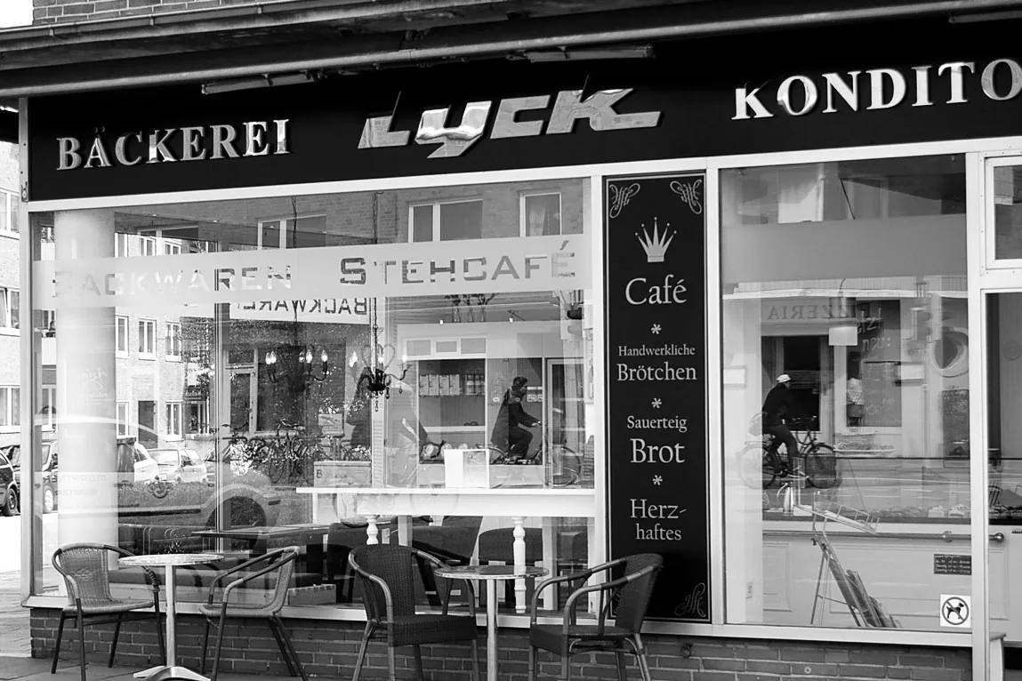 Die Filiale am Knooper Weg in einer Schwarzweißaufnahme: der Schriftzug „Bäckerei Lyck Konditorei“ über der Glasfront, davor Tische und Stühle des Stehcafés.