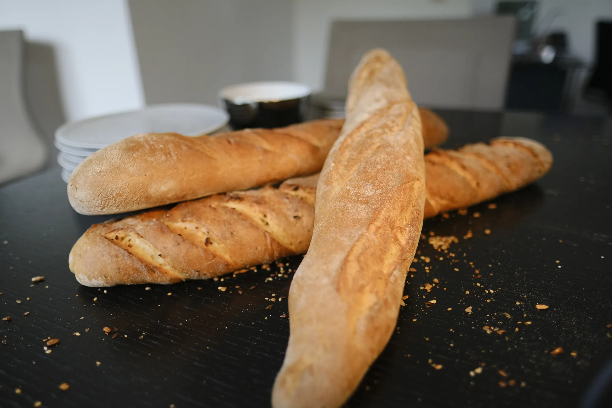 Vier Baguettes mit heller, aufgerissener Kruste liegen übereinander auf einem dunklen Tisch, dahinter gestapelte Teller.
