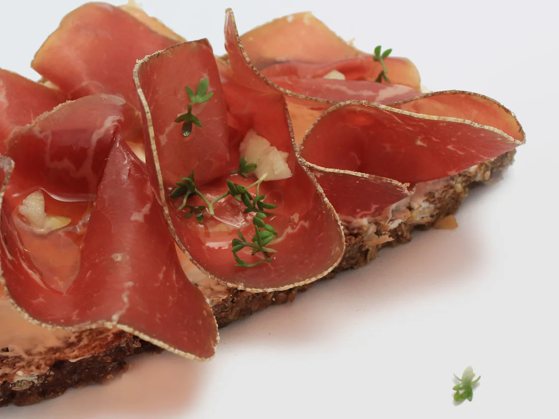 Dunkles Brot, dicht mit dünnen Scheiben Rauchfleisch belegt, dazwischen Thymian und Knoblauch.