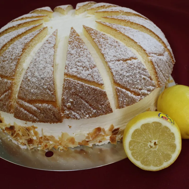 Eine gewölbte Torte mit Puderzucker, von hellen Sahnestreifen in Segmente geteilt, am Sockel Mandelblättchen; daneben eine ganze und eine halbierte Zitrone.